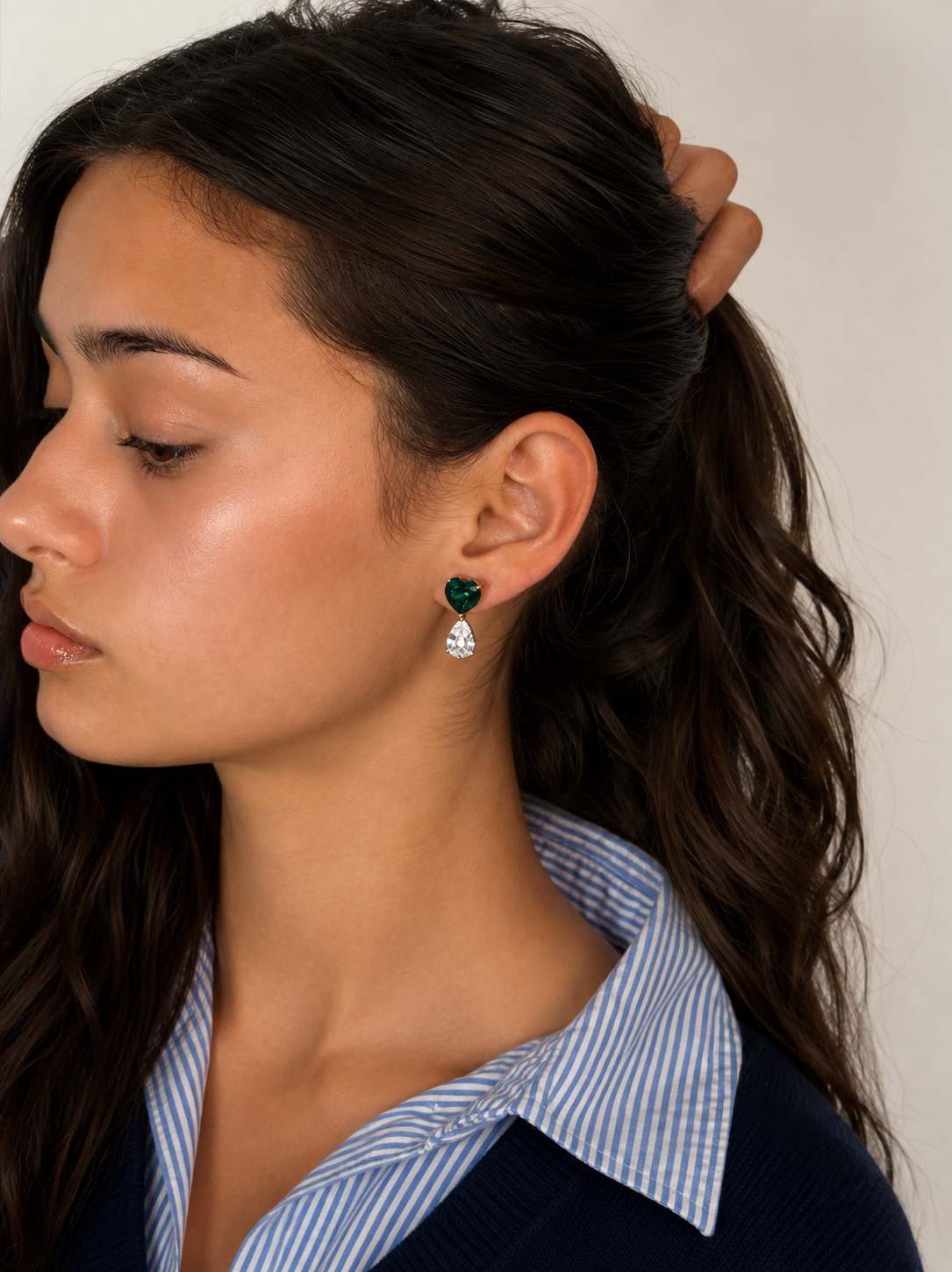 A close-up side profile of a woman with dark brown hair in a ponytail, wearing a green and clear teardrop dangle earring. She is wearing a blue and white striped collared shirt and a navy sweater, set against a plain light beige background.