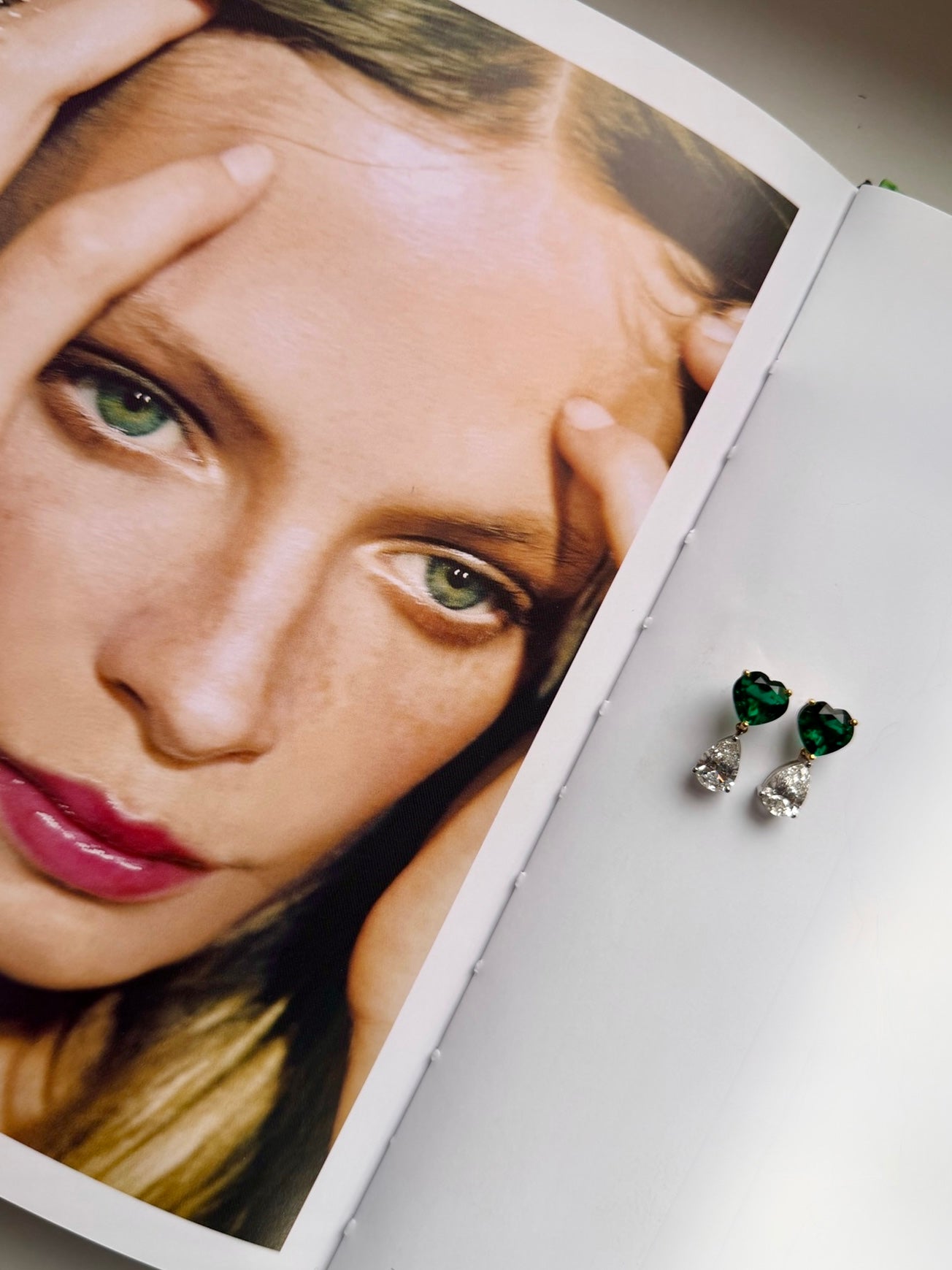 A pair of heart-shaped drop earrings with dark green stones and silver-toned clear crystal drops, resting on the edge of an open magazine featuring a close-up portrait.