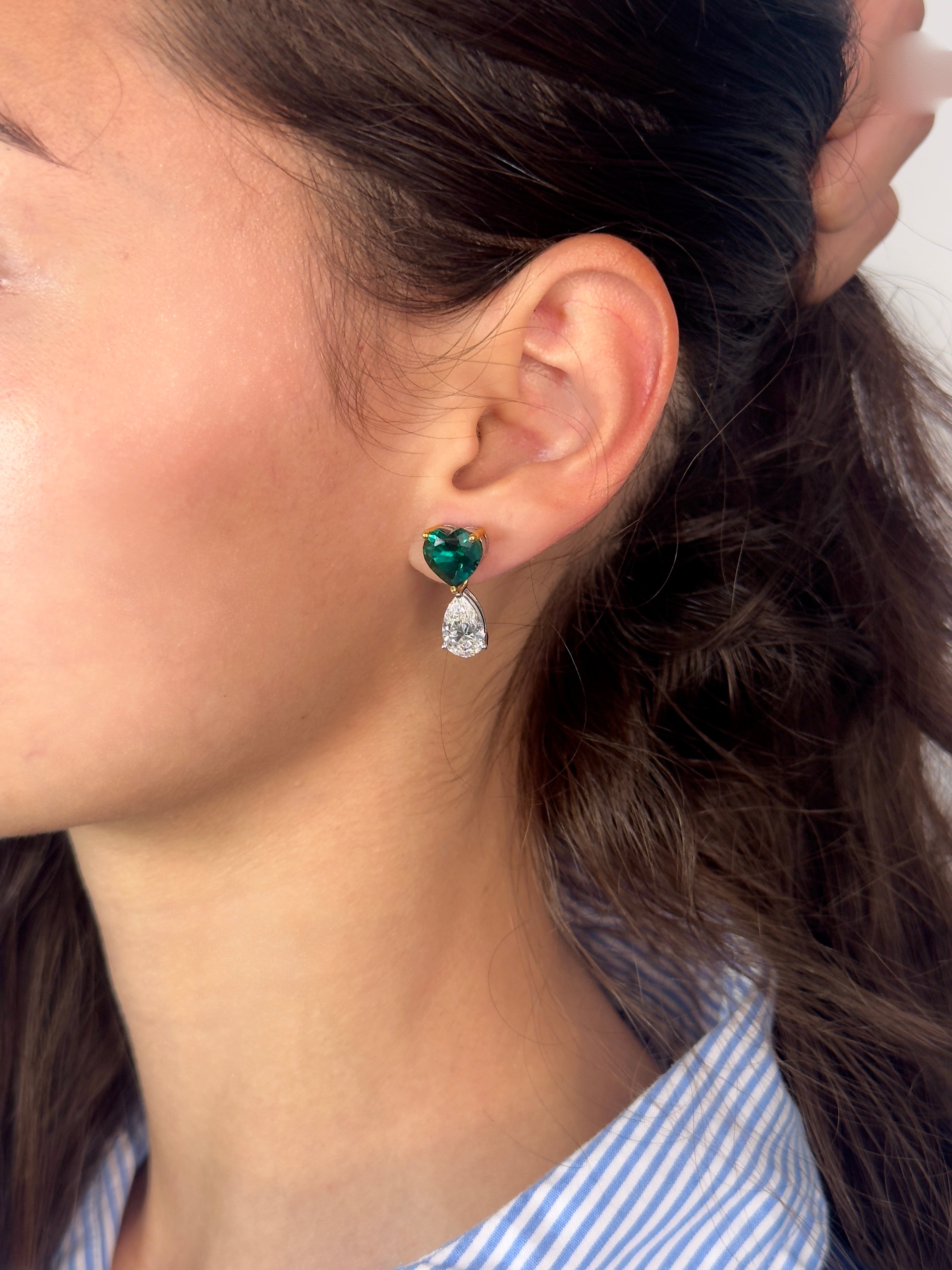 A close-up side profile of a woman wearing a small drop earring with a heart-shaped green gemstone and a teardrop-shaped clear stone below, set in a silver-toned metal.