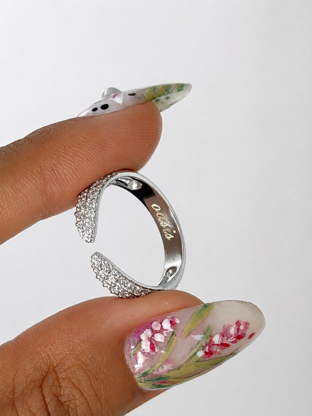 A silver open-bend ring with clear rhinestone accents on the curved ends, held between fingers with decorative floral nail art against a plain light background.
