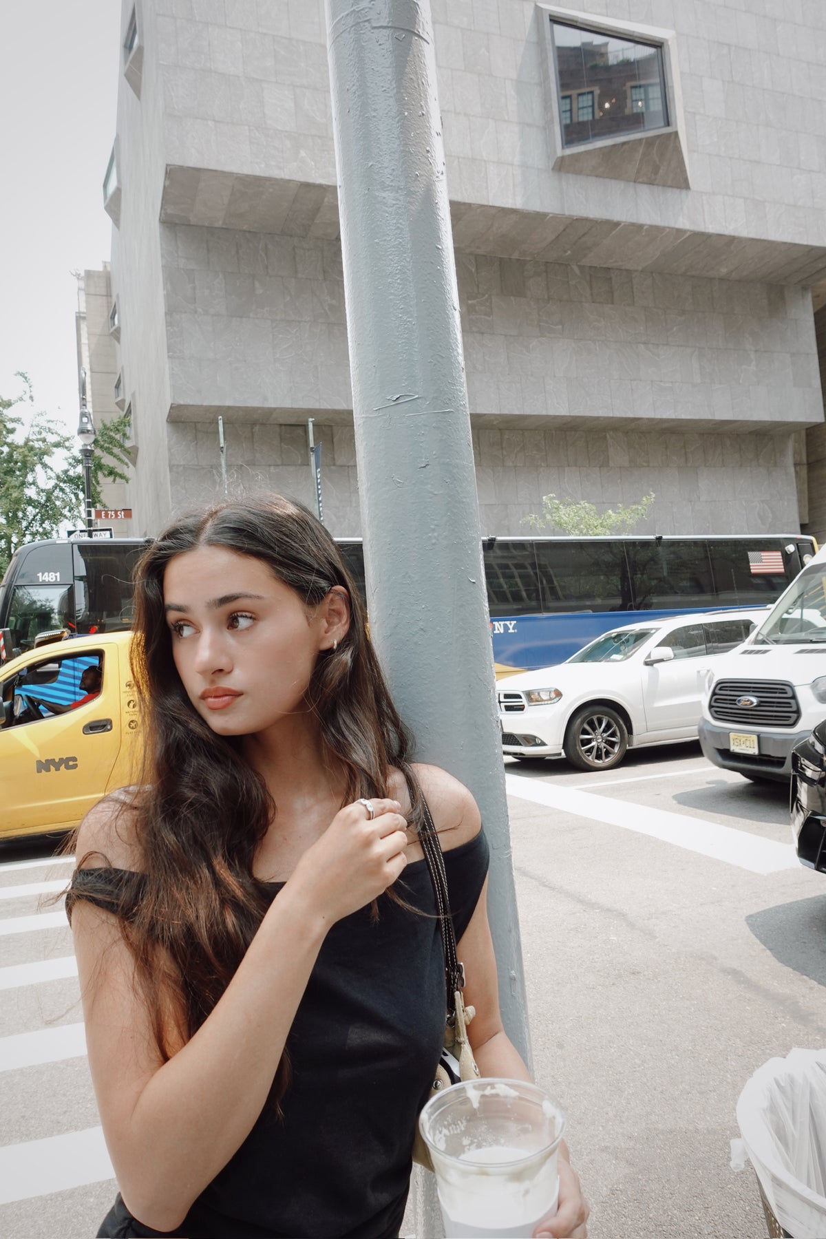 A woman with long dark hair stands on a city street beside a gray utility pole, wearing a black off-the-shoulder top and holding a clear plastic cup. Cars and a concrete building are visible in the background.
