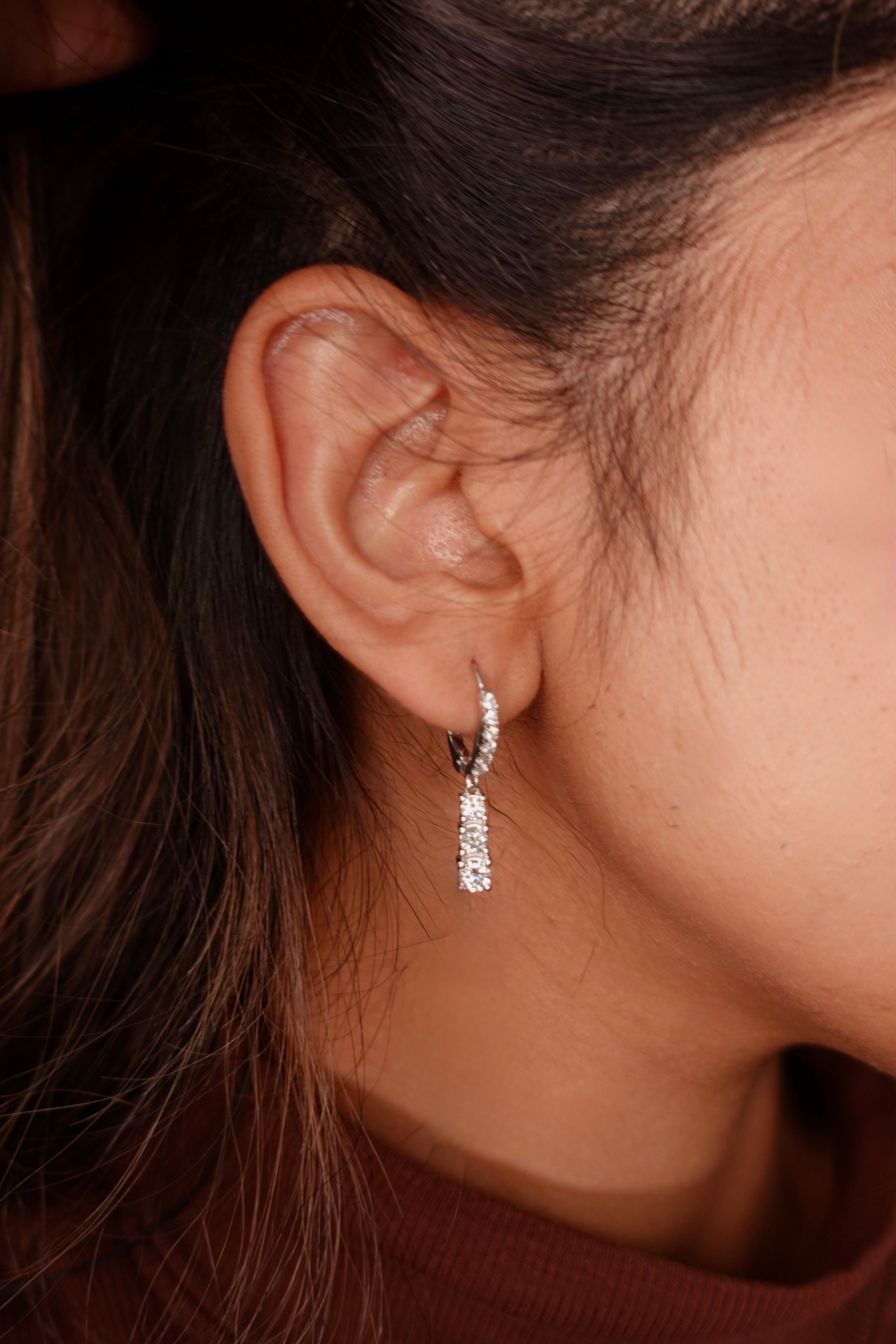 A close-up of a person's ear wearing a small silver hoop earring with a dangling clear stone-encrusted bar drop.