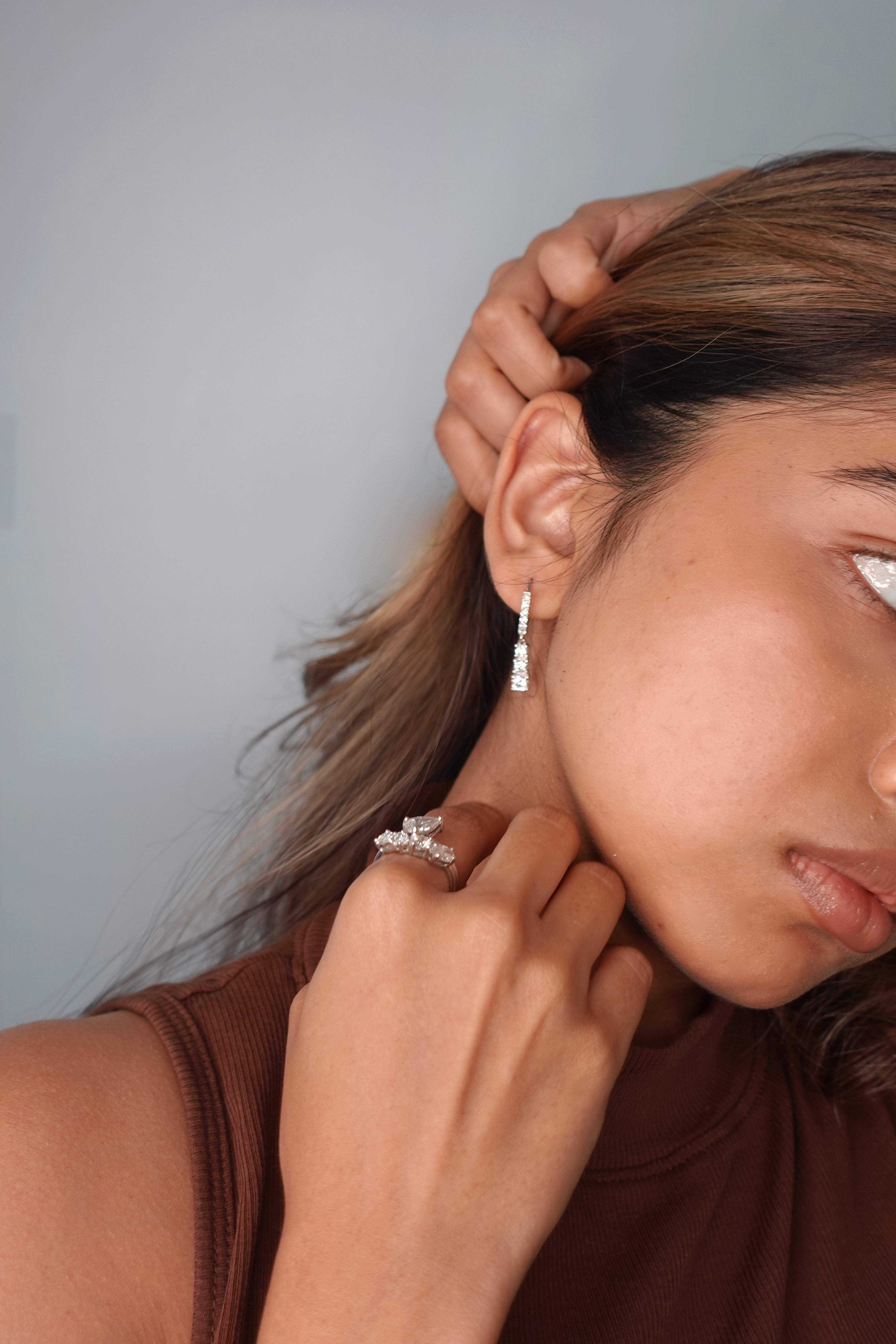 A close-up lifestyle shot of a woman wearing a delicate silver drop earring with a small clear gemstone and a silver ring on her finger, set against a plain light gray background.