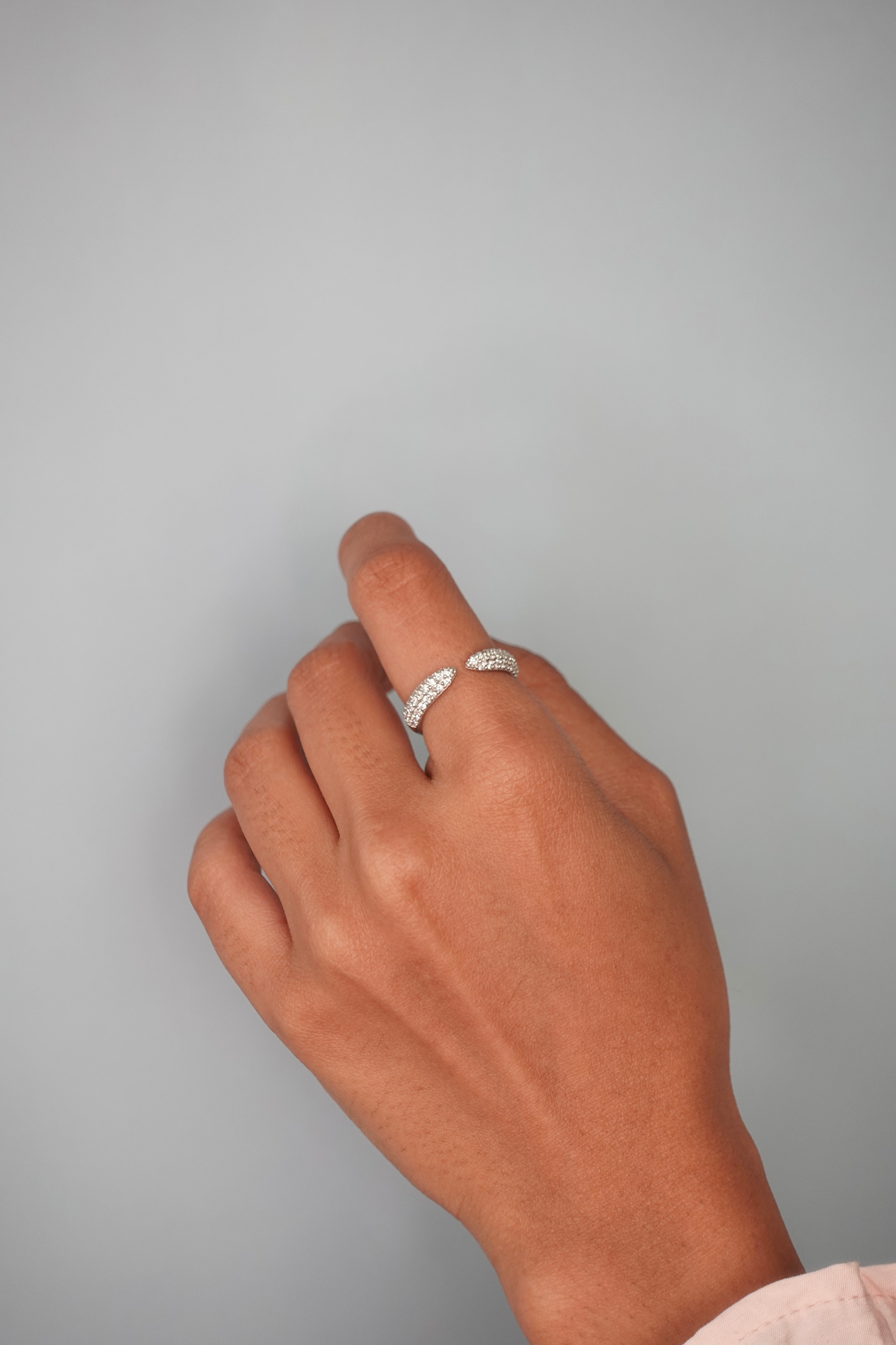 A close-up of a hand wearing a delicate silver ring with pavé-set clear stones, set against a solid light gray background.
