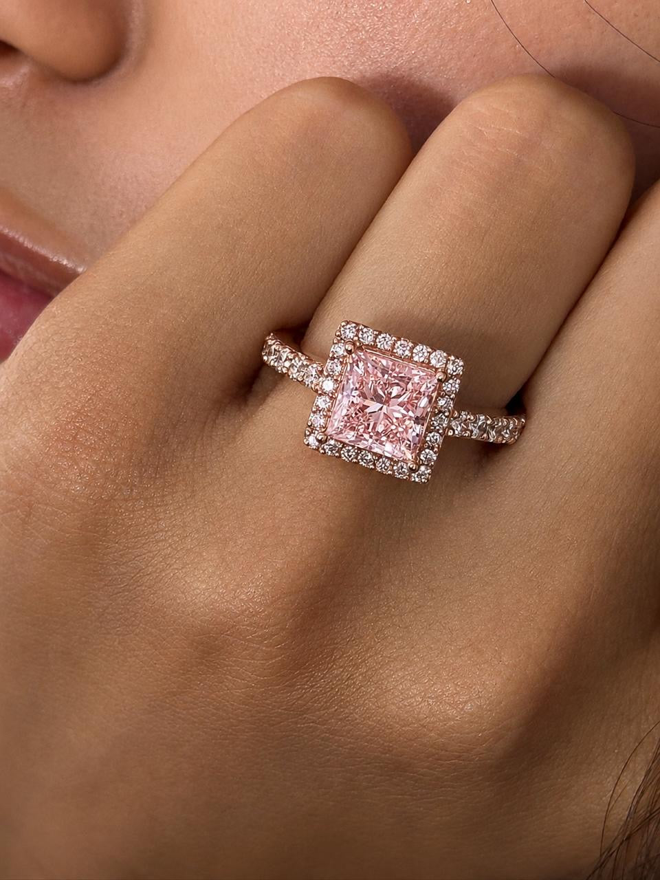 A close-up of a hand wearing a ring with a square pink gemstone surrounded by a halo of small clear stones, set in a rose gold-toned band with additional clear stones along the shoulders.