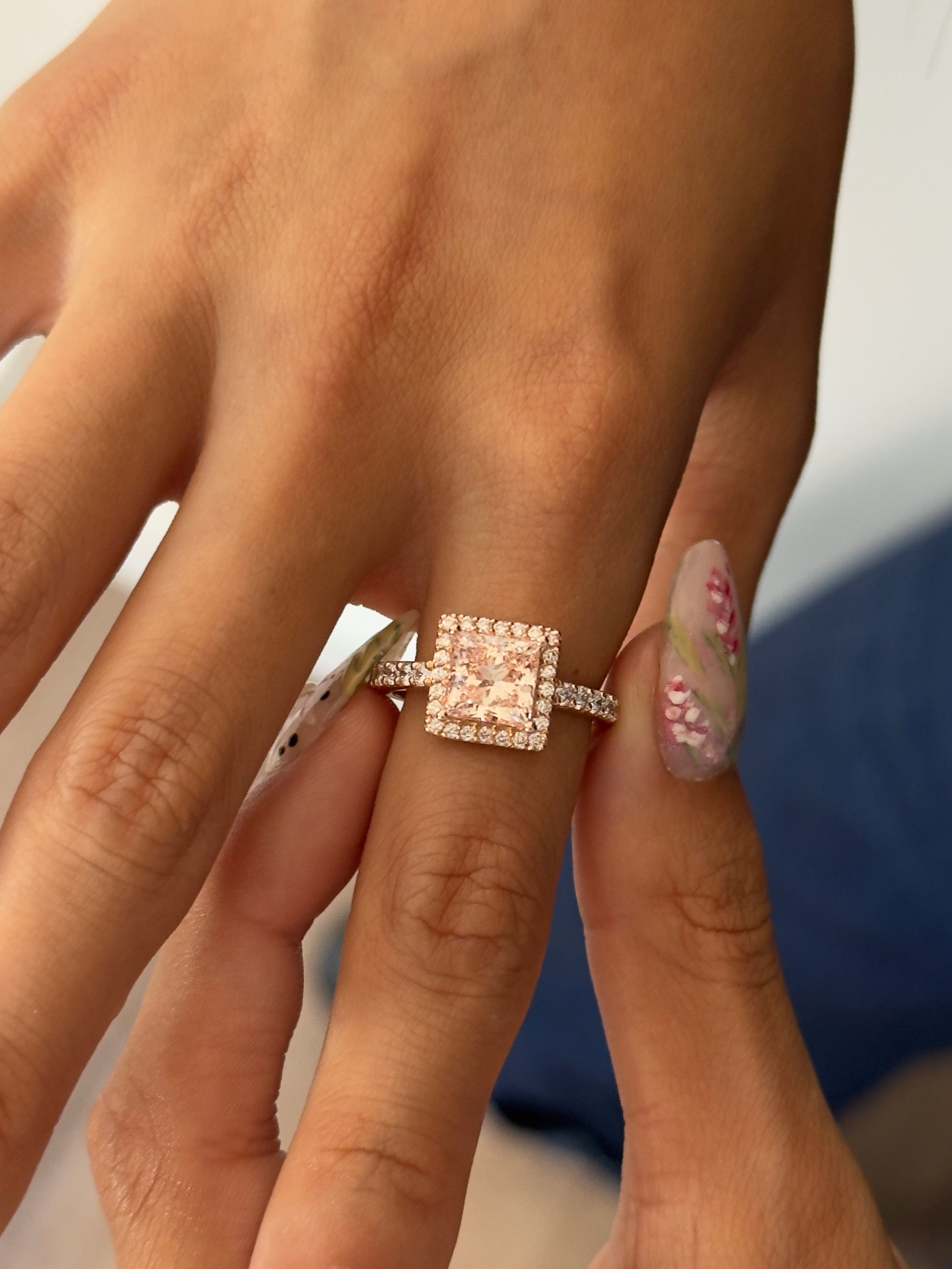 A close-up of a hand wearing a rose gold square halo ring with a central pink gemstone and small accent stones on the band.