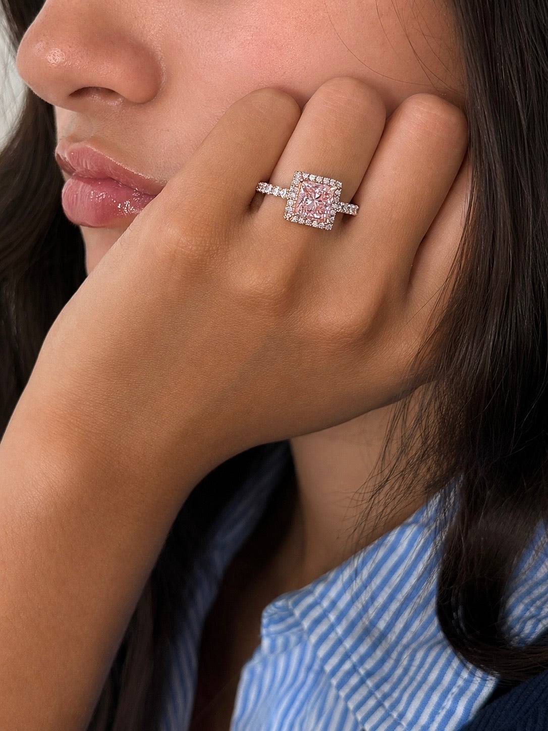 A close-up lifestyle shot of a woman wearing a pink diamond halo ring on her finger, resting her hand near her face.