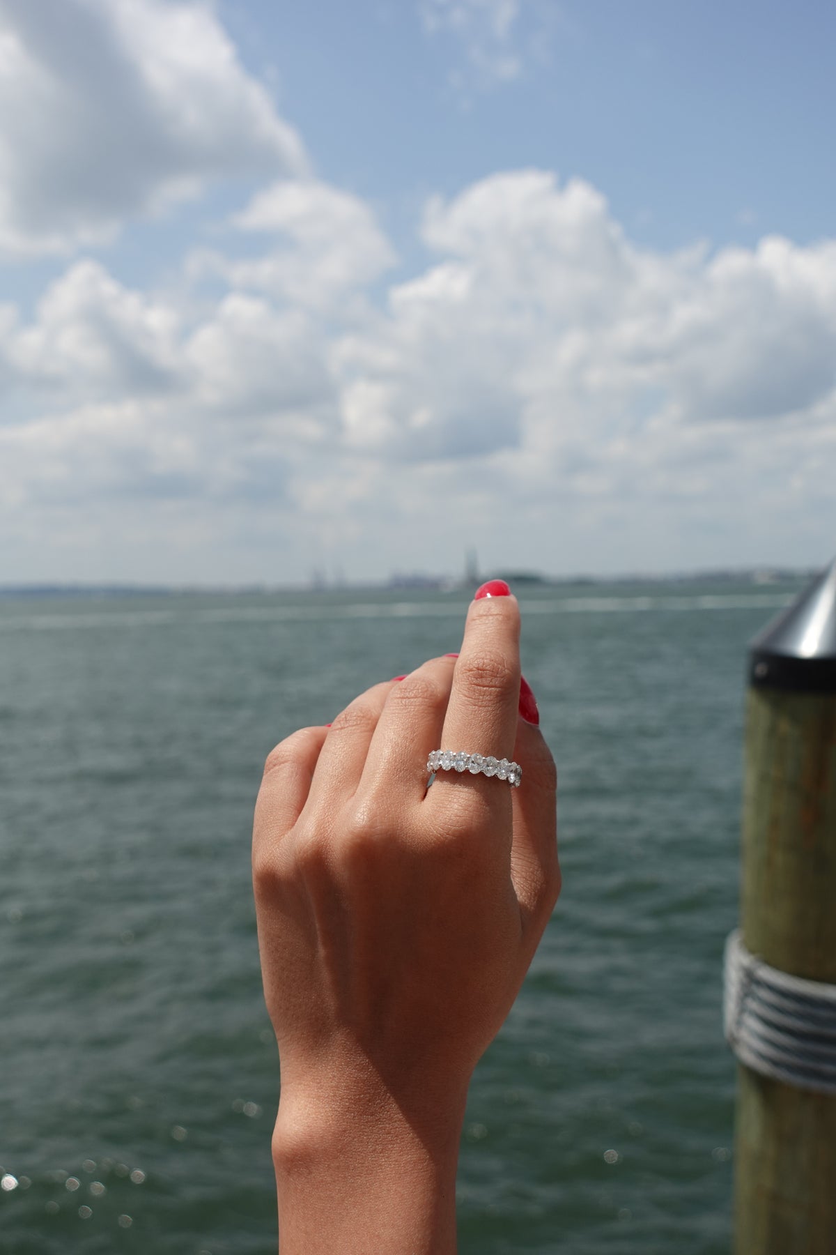 A close-up of a hand with red nail polish wearing a silver band ring with small clear stones, set against a blurred background of water and sky.
